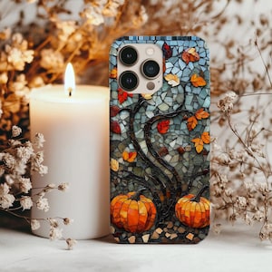 Op de afbeelding: Telefoonhoesje in mozaïekstijl met een boom met oranje pompoenen en herfstbladeren. De hoes heeft een blauw-grijze achtergrond en een zwarte rand. Een brandende witte kaars en droogbloemen staan op de achtergrond.