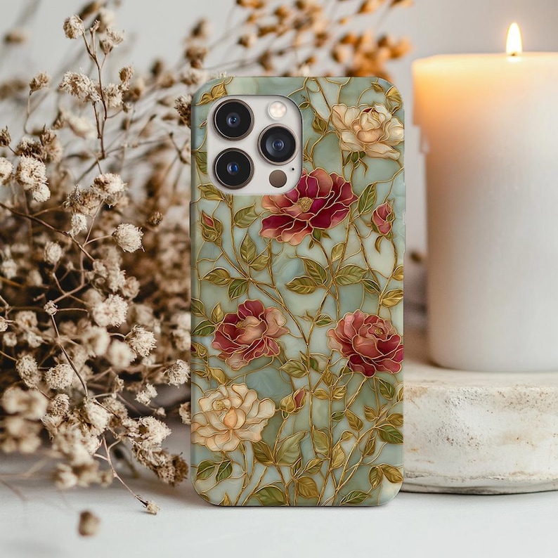 May include: A phone case featuring a floral design with red and cream-colored roses, green leaves, and gold-colored stems against a light blue background. The case has a glossy finish and a camera cutout.