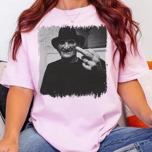 May include: A light pink t-shirt featuring a black and white image of a man in a hat giving the middle finger. The image has a distressed border. The man is wearing a black shirt.