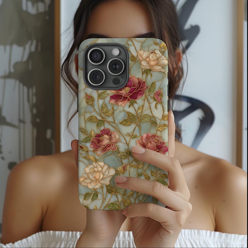 May include: A phone case featuring a floral design with red, pink, and cream-colored roses. The case has a light blue background with gold-colored vines and leaves. The phone case is held in front of a person.