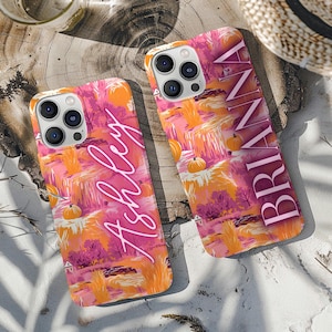 Op de afbeelding: Twee smartphonehoesjes met een levendige herfstlandschap-print met oranje pompoenen, roze bomen en gouden velden. Eén hoesje toont de naam Ashley in wit handschrift, terwijl de andere de naam BRIANNA in dikke witte letters weergeeft.