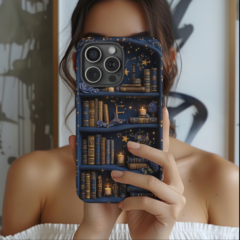May include: A dark blue phone case featuring a bookshelf design with miniature books, candles, and floral accents. The case has a starry night sky background with the letter "E" in the center. The case is held in front of a person.