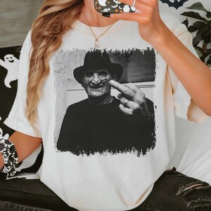 May include: Cream-colored t-shirt featuring a black and white image of a man in a hat, giving the middle finger. The graphic has a distressed border. The shirt is part of a Halloween-themed collection.
