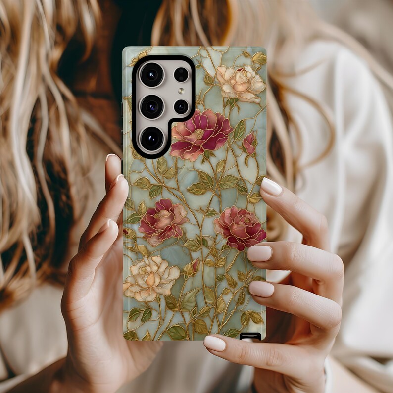 May include: A floral phone case featuring a design of burgundy and cream-colored roses with green leaves and gold stems against a light blue background. The case has a camera cutout and is held in a person's hands.