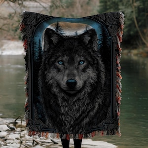 Op de afbeelding: Een geweven tapijtdeken met een gedetailleerde afbeelding van een zwarte wolf met doordringende blauwe ogen. De wolf staat tegen een achtergrond van een bos en een maanverlichte hemel. De deken heeft decoratieve franjes in rood en oranje.