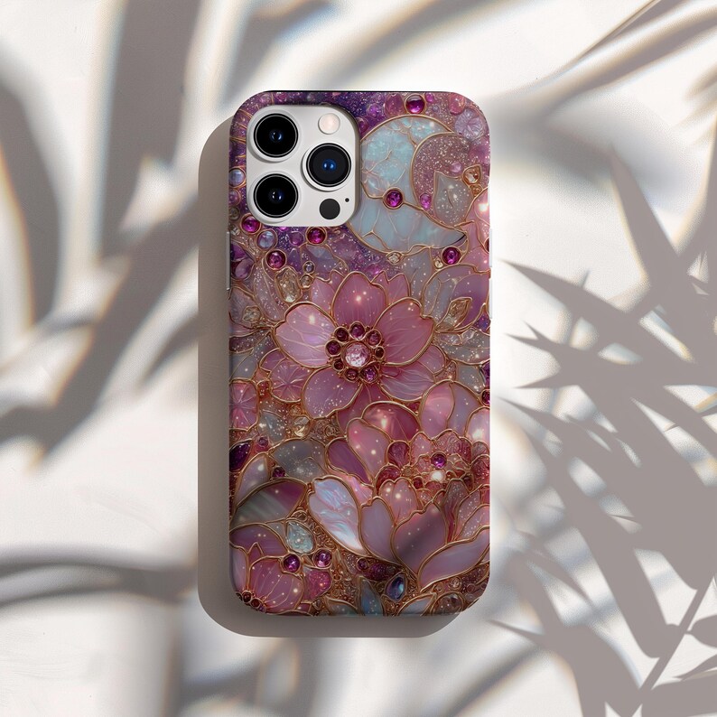 May include: A phone case with a floral design. The case features a pattern of pink and purple flowers with gold outlines and accents. Small, round, pink gems are scattered throughout the design. The case has a glossy finish.