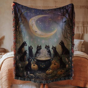 May include: A woven throw blanket featuring a whimsical Halloween scene. The design includes black cats gathered around a cauldron under a crescent moon, surrounded by pumpkins and trees. The blanket has a fringed edge.