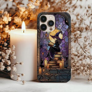 Puede incluir: Funda de teléfono con un diseño de vidriera que representa a una bruja leyendo sobre libros, con una luna llena de fondo. La funda tiene una combinación de colores púrpura y azul, con un gato negro y velas encendidas. Una vela blanca y flores secas están en el fondo.