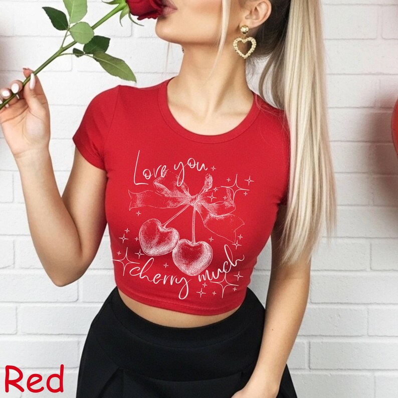 May include: A red crop top with a white graphic of two cherries, a bow, and the words "Love you" and "cherry much". The shirt has short sleeves and a round neckline. The word "Red" is at the bottom left.