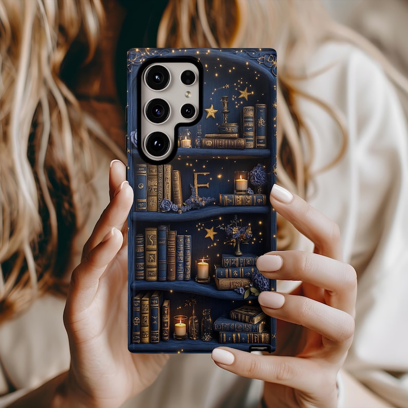 May include: A dark blue phone case featuring a bookshelf design with books, candles, and stars. The case has a detailed illustration with gold accents and the letter "F" on the shelves. The case is held by a person.