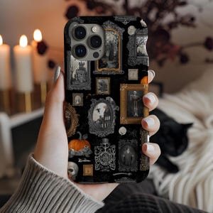 May include: Black phone case featuring a collage of framed vintage Halloween-themed illustrations. The artwork includes a skeleton, pumpkin, haunted house, and other spooky imagery. The case is held in a hand.