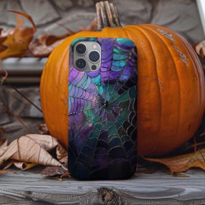 May include: A mobile phone case featuring a spiderweb design in purple, teal, and black. The case is placed in front of a large orange pumpkin and autumn leaves, creating a Halloween theme.