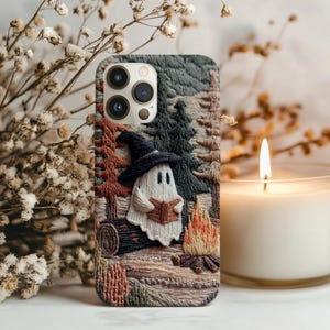 Puede incluir: Funda de teléfono bordada con una escena de acampada caprichosa con un fantasma con un sombrero de bruja, leyendo un libro junto a una fogata. La funda tiene un aspecto texturizado con un diseño de bosque y cabaña de madera, en tonos verdes, marrones y naranjas.