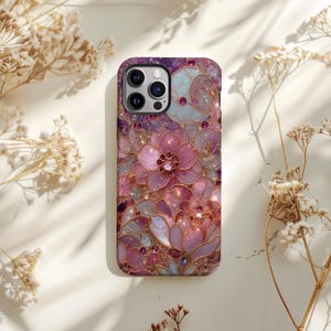 May include: A phone case with a floral design in shades of pink, purple, and gold. The case features a detailed pattern of flowers and leaves with jewel accents. The case has a glossy finish and a camera cutout.