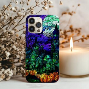 Könnte beinhalten: Eine Handyhülle zum Thema Halloween mit einem Buntglasdesign eines Spukhauses, Kürbissen und einem Vollmond. Die Hülle hat ein lila, grünes und orangefarbenes Farbschema. Die Handyhülle ist für ein Smartphone konzipiert.