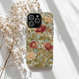 May include: A phone case featuring a floral design with red and cream roses, green leaves, and gold-colored stems on a light blue background. The case has a glossy finish and a camera cutout.