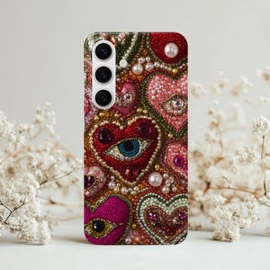 Puede incluir: Funda de teléfono decorativa con diseño de corazón y ojo, adornada con cuentas y perlas. La funda presenta una paleta de colores de rojo, rosa y verde. Se ve un recorte de cámara en la parte superior izquierda.