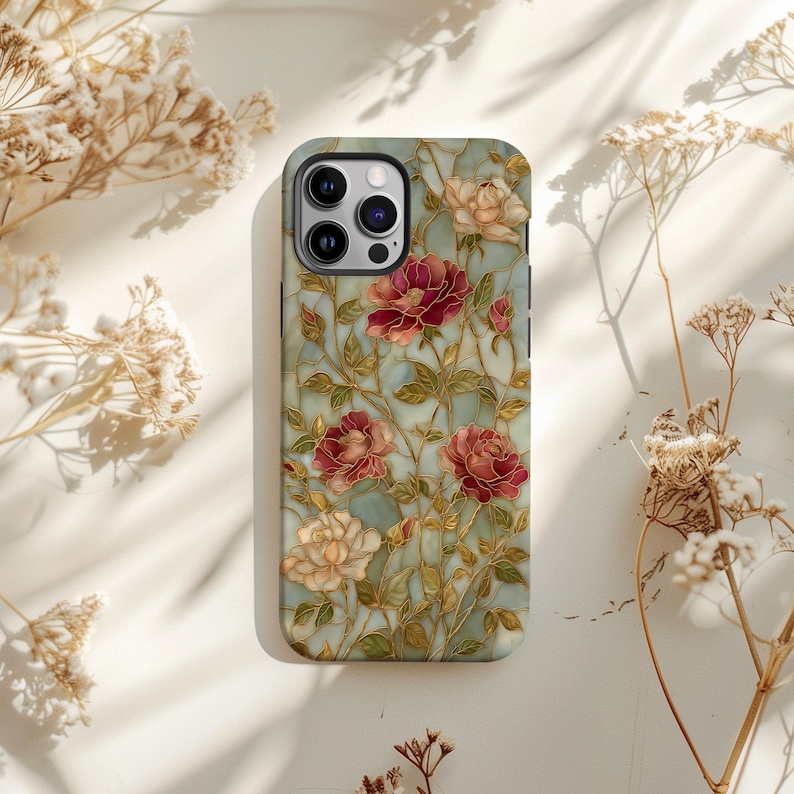 May include: A smartphone case featuring a floral design with red and cream-colored roses, green leaves, and gold-colored stems. The case has a light blue background and a glossy finish. The case has a camera cutout and buttons.