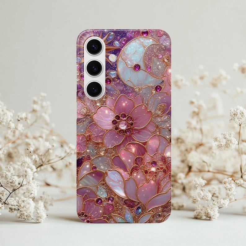 May include: A phone case featuring a floral design in shades of pink, purple, and gold. The case has a glossy finish and a detailed pattern of flowers, gems, and a crescent moon. The case has three camera lenses.