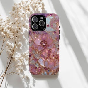 May include: A phone case with a floral design in shades of pink, purple, and gold. The case features a detailed pattern of flowers and leaves with sparkling accents. The camera lenses are visible at the top.