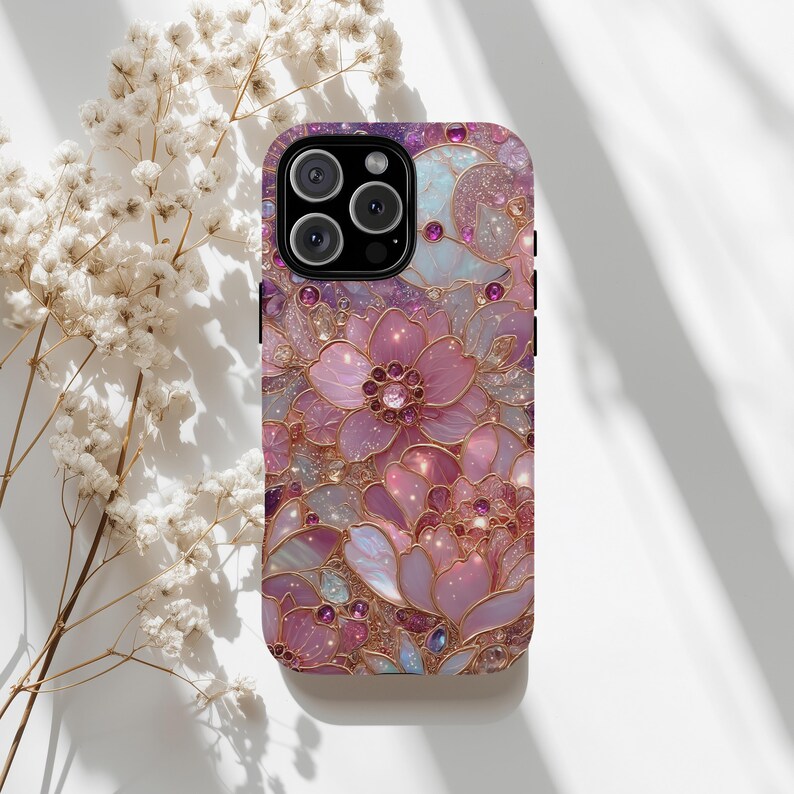 May include: A phone case with a floral design in shades of pink, purple, and gold. The case features a detailed pattern of flowers and leaves with sparkling accents. The camera lenses are visible at the top.