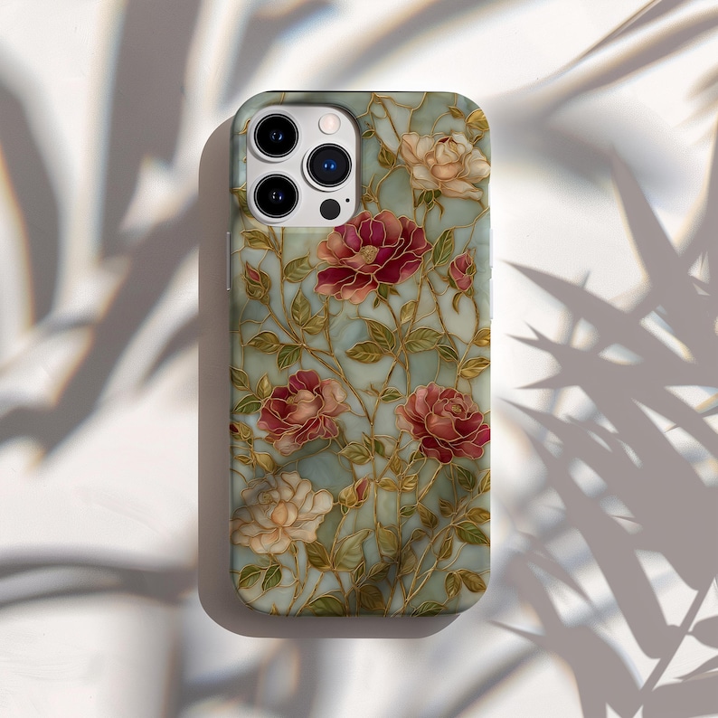 May include: A phone case featuring a floral design with red and cream-colored roses, green leaves, and gold-colored stems against a light blue background. The case has a glossy finish and a camera cutout.