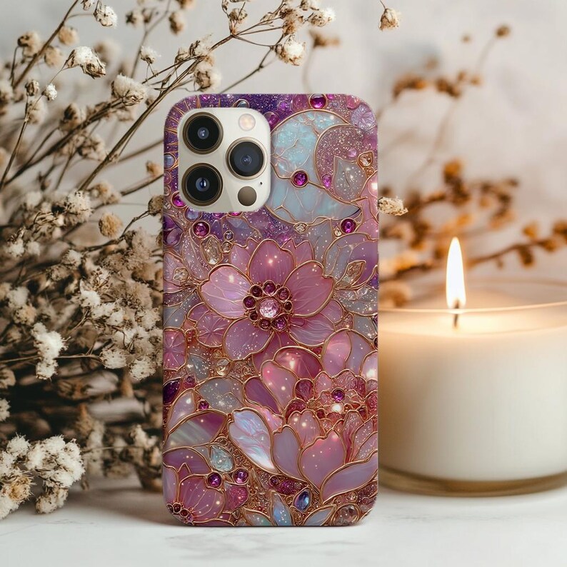 May include: A phone case with a floral design in shades of purple, pink, and gold. The case features a detailed pattern of flowers and gemstones. The phone case is shown with a lit candle and white flowers in the background.