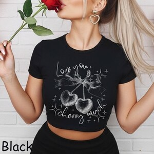 May include: Black crop top with a white graphic of two cherries tied with a bow and the words "Love you cherry much". The shirt has short sleeves and a round neckline. The word "Black" is printed at the bottom left.