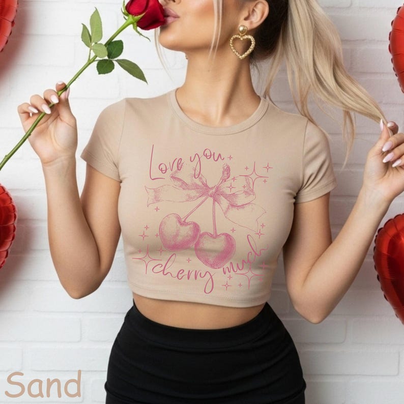 May include: Sand-colored crop top with a pink cherry graphic and the text "Love you cherry much." The top has short sleeves and a round neckline. The model is holding a red rose.