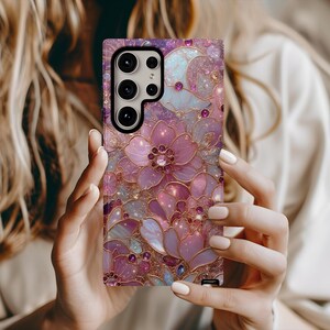 May include: A phone case with a floral design in shades of pink, purple, and gold. The case features a detailed pattern of flowers and leaves with a sparkling, glittery effect. The case has a camera cutout and buttons.