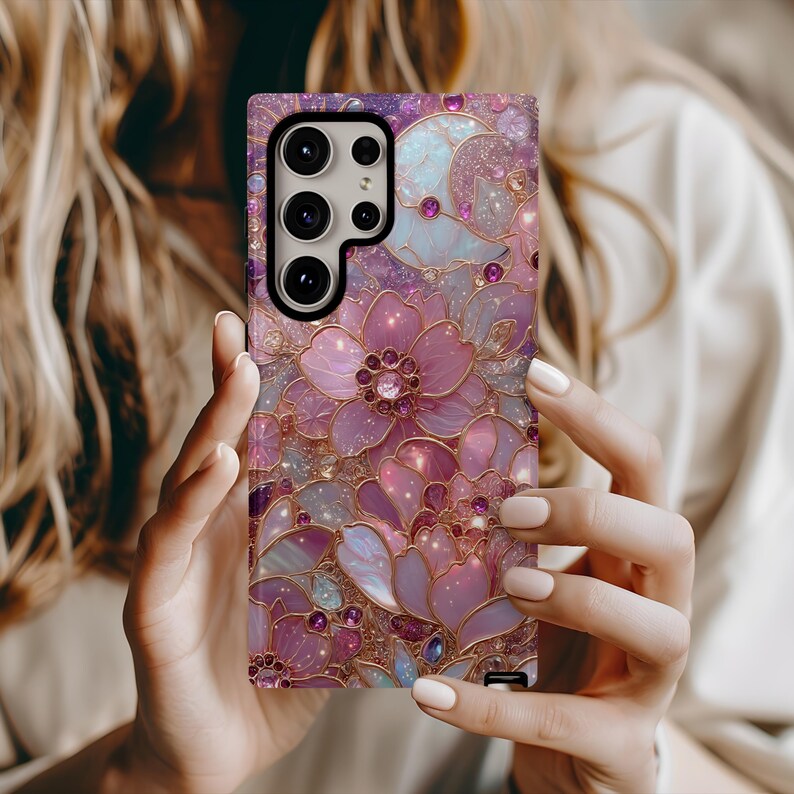 May include: A phone case with a floral design in shades of pink, purple, and gold. The case features a detailed pattern of flowers and leaves with a sparkling, glittery effect. The case has a camera cutout and buttons.