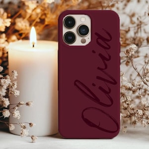 May include: A burgundy phone case with the name "Olivia" in a cursive font. The case has a triple-lens camera cutout. A lit white candle and dried flowers are in the background.