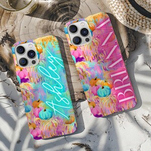 Op de afbeelding: Twee smartphonehoesjes met een levendig, abstract, veelkleurig patroon van pompoenen en hooi. Het ene hoesje toont de naam ASHLEY in een helder cyaan handschrift, terwijl het andere de naam BRIANNA in een vet, roze hoofdlettertype heeft.
