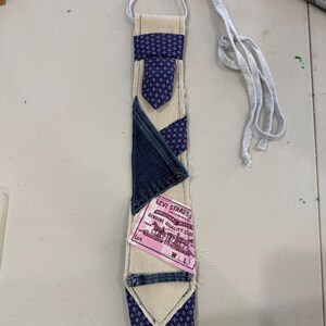 May include: A necktie crafted from a patchwork of cream-colored canvas, dark blue denim, and a purple fabric with a small white geometric pattern. A pink Levi Strauss brand label is stitched onto the center, featuring text and horse imagery.