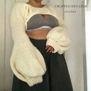 Puede incluir: Un suéter blanco de punto de ganchillo recortado con mangas largas y abullonadas. El suéter tiene un corte en la parte delantera, dejando ver la piel debajo. El texto "CROPPED SWEATER crochet" está escrito en negro en la parte superior de la imagen.
