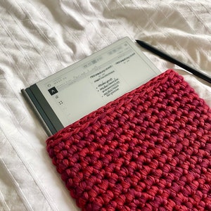 Peut inclure: Un lecteur électronique blanc avec une couverture noire est posé sur un couvre-lit blanc. Le lecteur électronique est partiellement recouvert d'une pochette en crochet rouge. L'écran du lecteur électronique affiche une liste de tâches manuscrites.