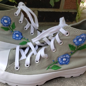Puede incluir: Un par de zapatillas de lona de caña alta con cordones blancos y flores azules bordadas con tallos verdes sobre un fondo verde salvia.
