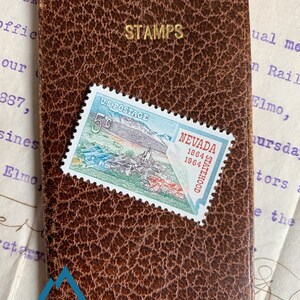 K&ouml;nnte beinhalten: Ein braunes Leder-Briefmarkenalbum mit dem in Gold gepr&auml;gten Wort "STAMPS". Eine 5-Cent-US-Briefmarke mit einer malerischen Ansicht von Nevada ist auf dem Album angebracht. Die Marke zeigt eine Bergkette, eine Stadt und den Text "NEVADA 1864 1964 STATEHOOD".