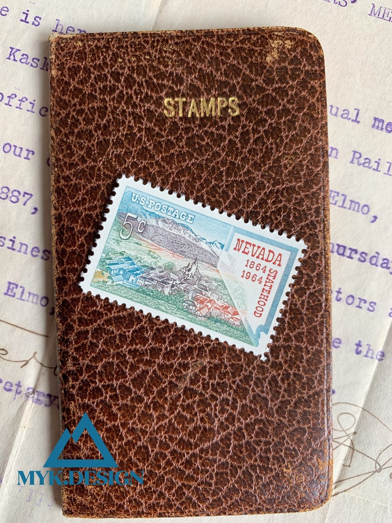K&ouml;nnte beinhalten: Ein braunes Leder-Briefmarkenalbum mit dem in Gold gepr&auml;gten Wort "STAMPS". Eine 5-Cent-US-Briefmarke mit einer malerischen Ansicht von Nevada ist auf dem Album angebracht. Die Marke zeigt eine Bergkette, eine Stadt und den Text "NEVADA 1864 1964 STATEHOOD".