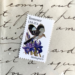 May include: A vintage postage stamp featuring a brown and white mockingbird perched on a purple iris. The stamp is from Tennessee and reads "USA 20c".
