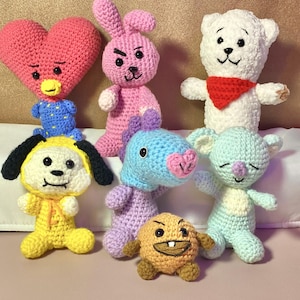 May include: A collection of seven handmade crochet dolls in various colors. The dolls include a pink heart, a pink bunny, a white bear with a red scarf, a yellow dog, a blue dinosaur, a light blue koala, and a brown round character.