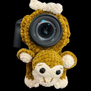 May include: A mustard yellow crochet camera lens cover in the shape of a monkey. The monkey has white detailing on its ears, face, and paws, with black button eyes. The camera lens is visible in the centre of the monkey's face.