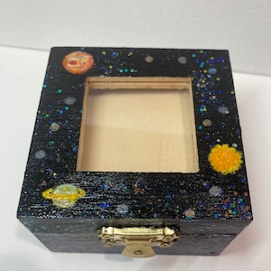 May include: A small wooden box with a black background and glitter. The box has a clear window and a latch. The box is decorated with painted planets and stars.