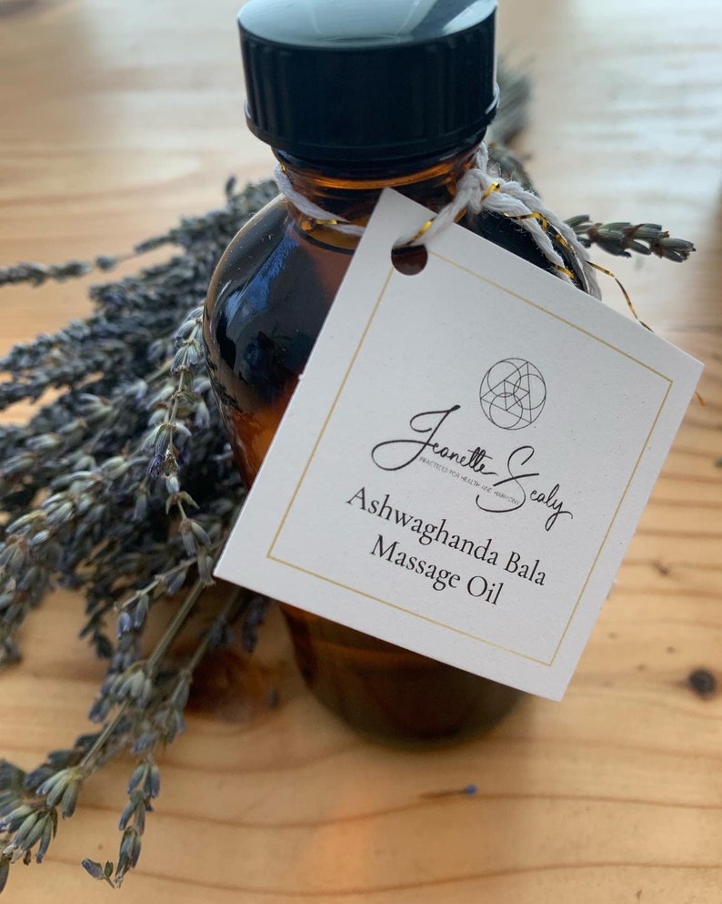 May include: A small amber glass bottle with a black lid and a white label. The label has a gold circle with a geometric design and the text "Jeanette Sealy PRACTICED FOR HEALTH & WELLNESS" and "Ashwagandha Bala Massage Oil". The bottle is sitting on a wooden surface with dried lavender flowers.