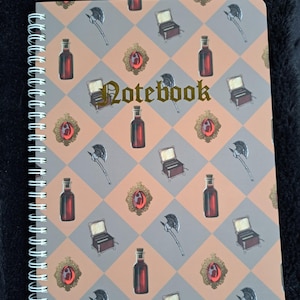May include: Spiral-bound notebook with a repeating pattern of fantasy-themed illustrations. The design includes bottles, axes, treasure chests, and ornate frames with red gems, set against a peach and gray checkered background. The word "Notebook" is written in gold script.