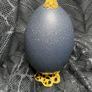 May include: A dark blue decorative egg with a textured surface, topped with a gold-colored floral embellishment. The egg rests on a gold-colored filigree base, creating an elegant display.
