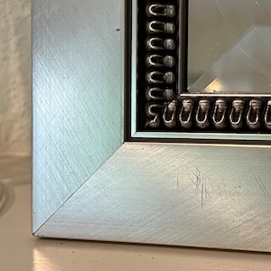 May include: Close-up of the corner of a silver picture frame with a black and silver decorative border. The frame has a few scratches on the surface.