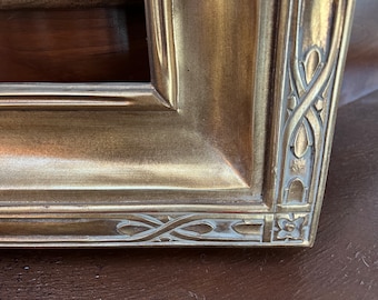 May include: Close-up of a gold picture frame with an ornate design. The frame has a raised edge and a detailed pattern around the perimeter.