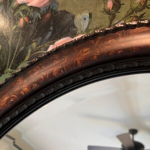 May include: A close-up of an oval mirror with a dark brown wood frame. The frame has a decorative, swirling pattern in a lighter brown color.