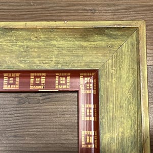 May include: Close-up of a wooden picture frame with a green and red color scheme. The frame has a rectangular shape with a decorative pattern in the red section. The wood grain is visible.